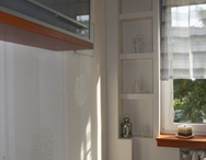 Ściana z zamontowanymi płytami gipsowo-kartonowymi, tworzącymi wnęki dekoracyjne, oświetlona naturalnym światłem wpadającym przez okno z roletą rzymską.