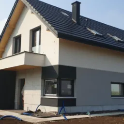 Nowoczesny dom jednorodzinny z elewacją w odcieniach bieli i szarości, czarną stolarką okienną i dachem z dachówki. Widoczne przygotowania do zagospodarowania terenu.