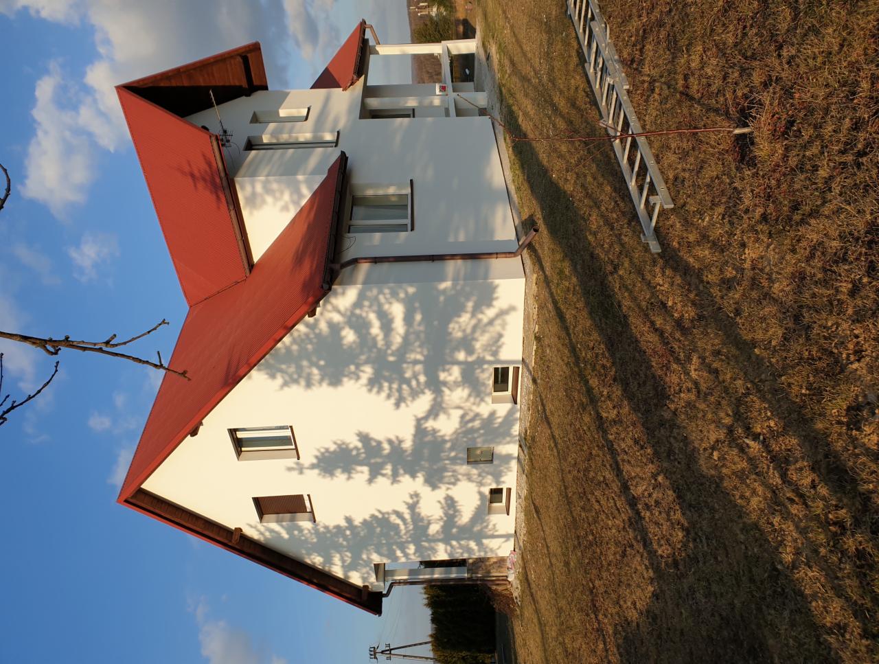 Nowoczesny dom z czerwoną dachówką i białą elewacją, cień drzewa na ścianie, drabina oparta o ścianę, widok z boku w słoneczny dzień.