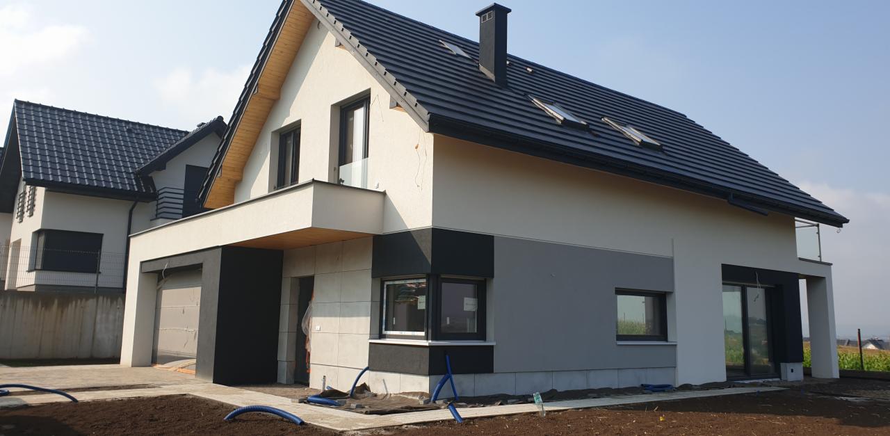 Nowoczesny dom jednorodzinny z elewacją w odcieniach bieli i szarości, czarną stolarką okienną i dachem z dachówki. Widoczne przygotowania do zagospodarowania terenu.