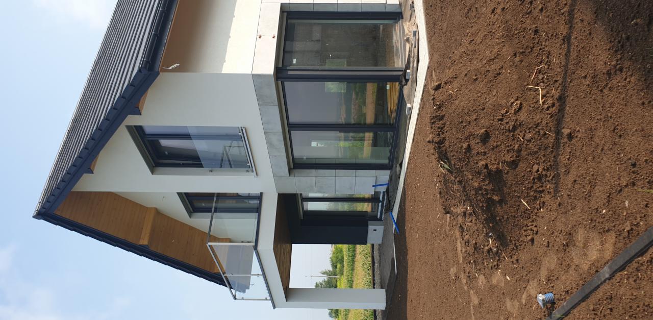 Nowoczesny dom z elewacją w odcieniach bieli i szarości, z dużymi oknami i szklaną balustradą balkonu. Widoczny fragment przygotowanego terenu.