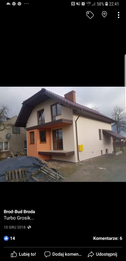 Dwukondygnacyjny dom jednorodzinny z częściowo pomalowaną elewacją w odcieniach beżu i pomarańczy, brązowe okna i dach, widoczny stos materiałów budowlanych na pierwszym planie.