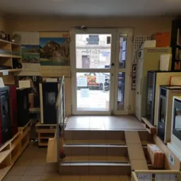 Wnętrze sklepu z ekspozycją pieców na pellet na drewnianych paletach, schody prowadzące do wejścia, regał z akcesoriami i półka z książkami w tle.