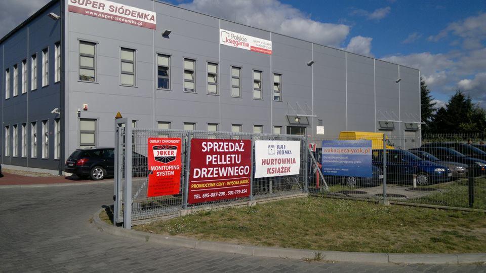 Szary budynek magazynowy z reklamami na ogrodzeniu, w tym ogłoszenie sprzedaży pelletu drzewnego.