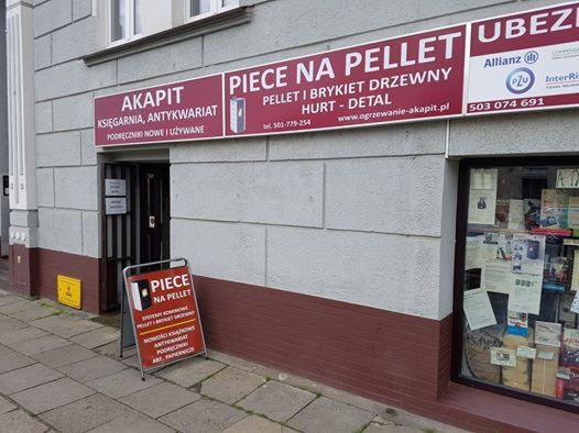 Fasada budynku z szyldem 'Akapit Księgarnia, Antykwariat' obok reklamy 'Piece na pellet, pellet i brykiet drzewny, hurt-detal' oraz szyldem ubezpieczeń Allianz i PZU, dodatkowo na chodniku stoi...