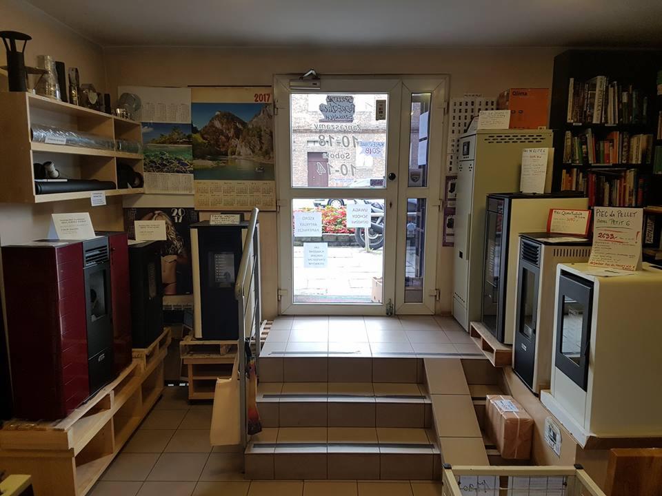 Wnętrze sklepu z ekspozycją pieców na pellet na drewnianych paletach, schody prowadzące do wejścia, regał z akcesoriami i półka z książkami w tle.
