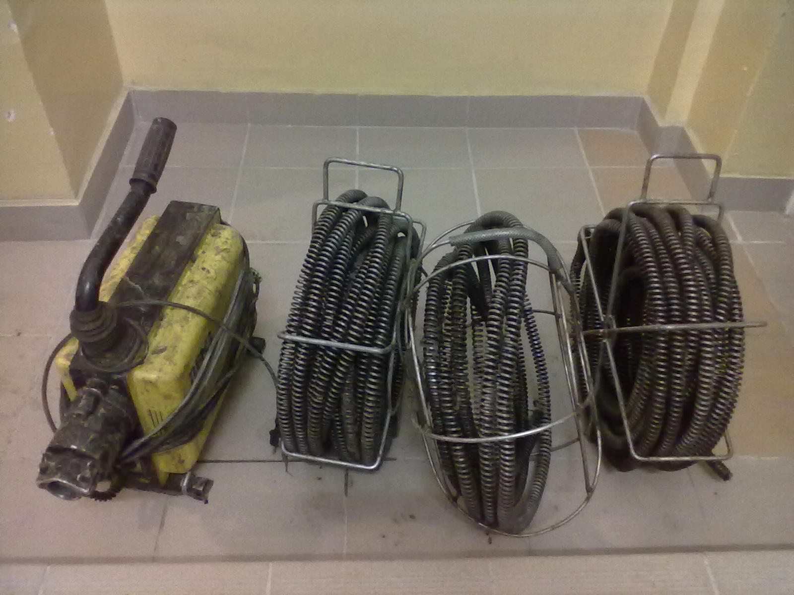 Elektryczna przepychaczka do rur kanalizacyjnych z trzema bębnami ze sprężyną spiralną w stalowych koszach, stojąca na kafelkach pod ścianą.