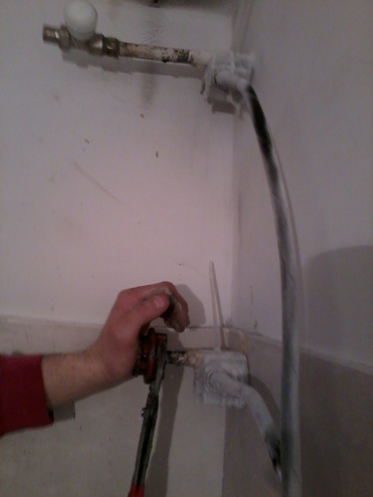 Ręka hydraulika zaciska zamarzniętą rurę narzędziem do gwintowania rur w rogu pomieszczenia.