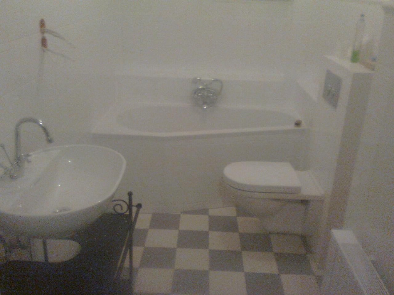 Jasna łazienka z białą wanną, toaletą i umywalką na ozdobnym stelażu, wykończona szachownicą z płytek. Minimalistyczny design, świeżo po remoncie.