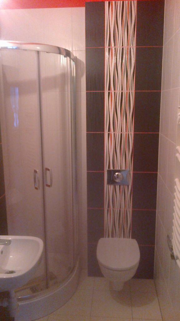 Wykończona łazienka z kabiną prysznicową, umywalką i toaletą. Na ścianie dekoracyjne płytki w odcieniach brązu i bieli z czerwonymi akcentami. Widoczny grzejnik.