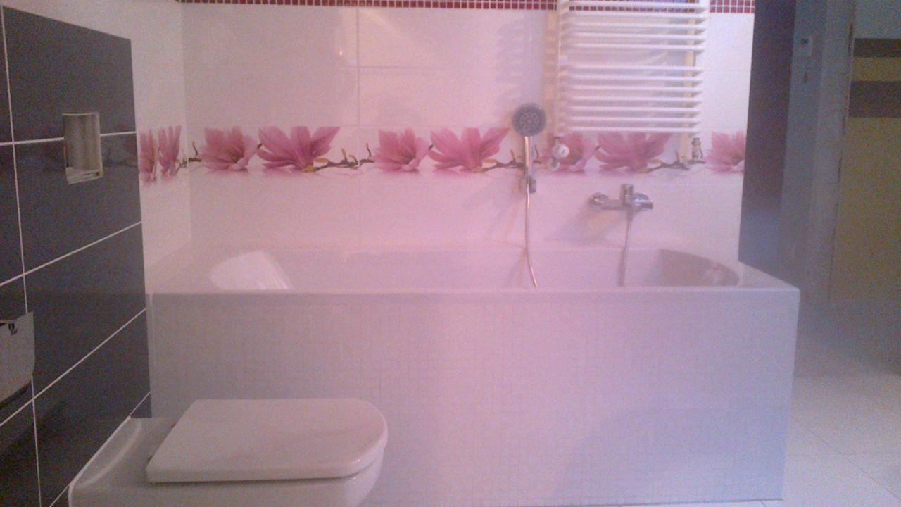 Wykończona łazienka z białą wanną obudowaną płytkami, toaleta i grzejnik drabinkowy. Na ścianach płytki z motywem kwiatowym i ciemne płytki po lewej stronie.