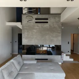 Nowoczesny salon z szarą narożną sofą, kominkiem obudowanym płytami imitującymi beton i wiszącą lampą pierścieniową, oświetlenie punktowe w suficie, widoczna podłoga z paneli.