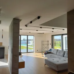 Nowoczesne wnętrze domu z dużymi oknami przesuwnymi wychodzącymi na basen, drewniana podłoga, szara sofa modułowa i dekoracyjny filar z efektem betonu.