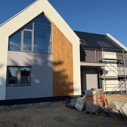 Usługi Remontowo-Budowlane DECORMANN - Marcin Wocheń - Nowoczesny dom jednorodzinny z elewacją w odcieniach szarości i beżu, z elementami drewnianymi i rusztowaniem. Widoczne materiały budowlane.