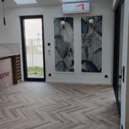 Parkiet ułożony we wzór jodełki francuskiej w jasnym wnętrzu z oknami dachowymi, klimatyzacją w folii i dekoracyjnymi lampami ściennymi.