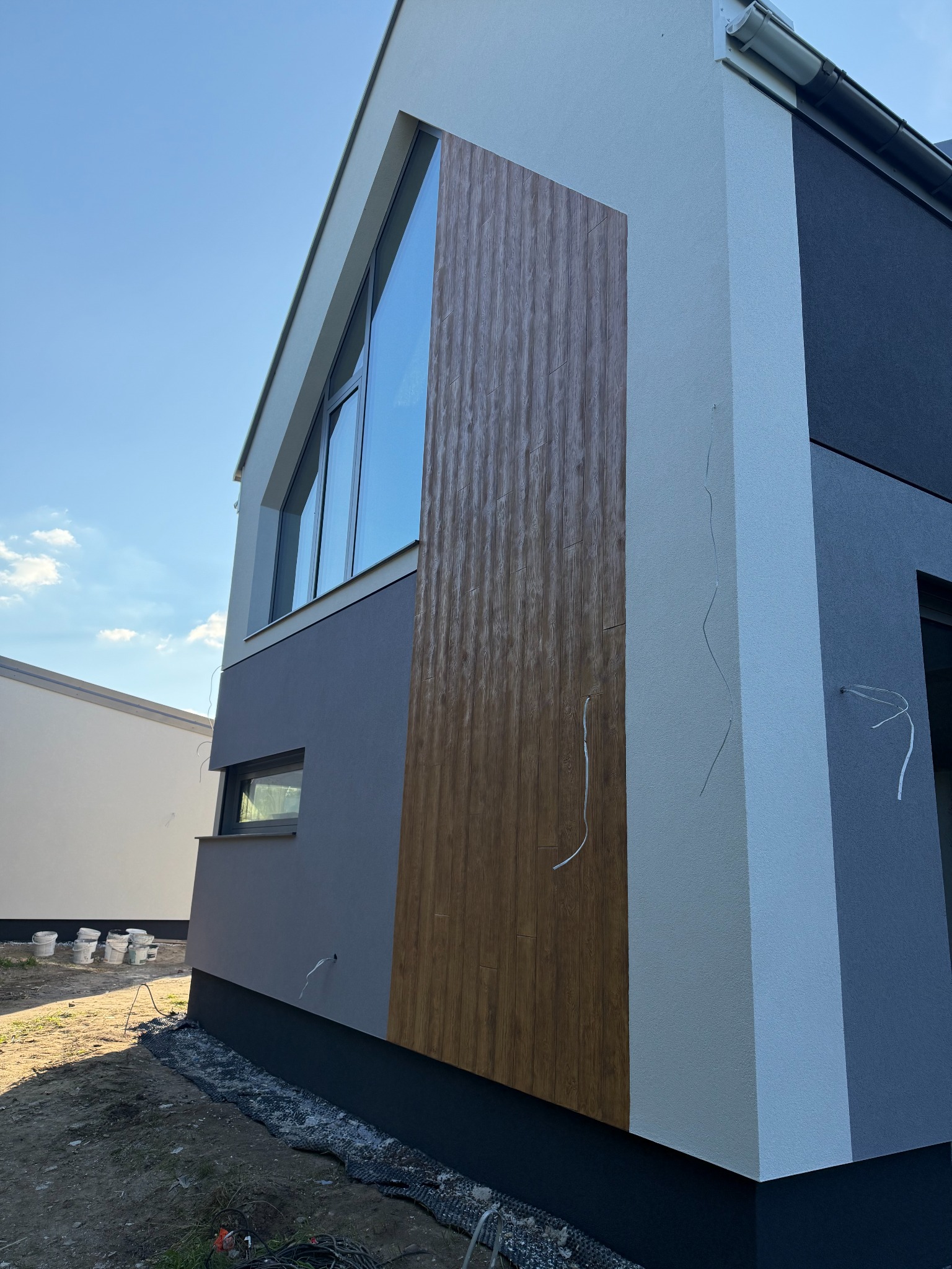 Fragment elewacji domu z drewnianą okładziną, widoczne puszki po farbie i przewody. Nowoczesne wykończenie budynku w odcieniach szarości i bieli pod błękitnym niebem.