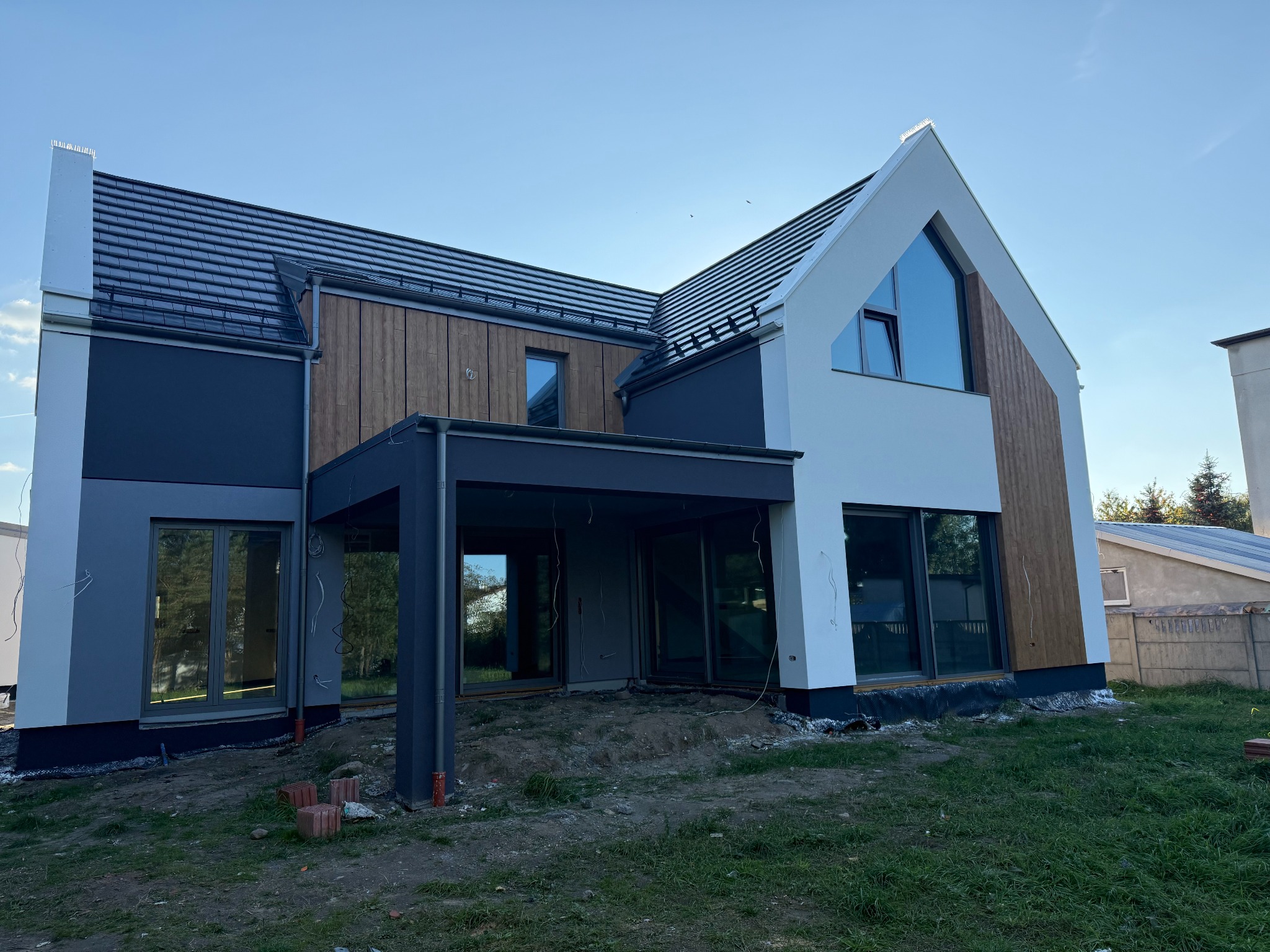 Nowoczesny dom jednorodzinny z elementami drewnianej elewacji i dużymi oknami, widziany z zewnątrz w słoneczny dzień, w trakcie prac wykończeniowych.