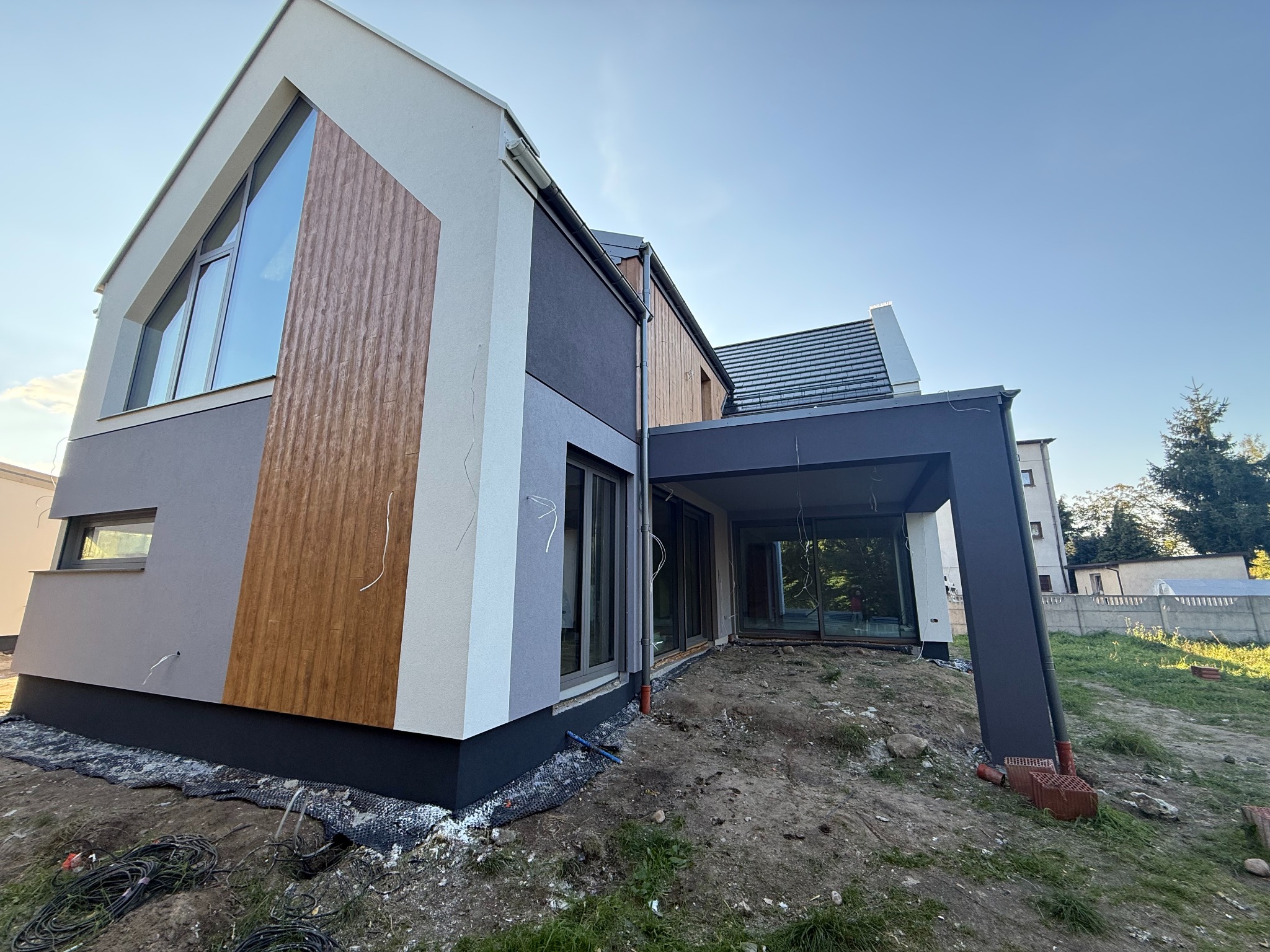 Nowoczesny dom jednorodzinny z elementami drewnianymi na elewacji, duże okna, antracytowa podmurówka, widoczne prace wykończeniowe wokół budynku.