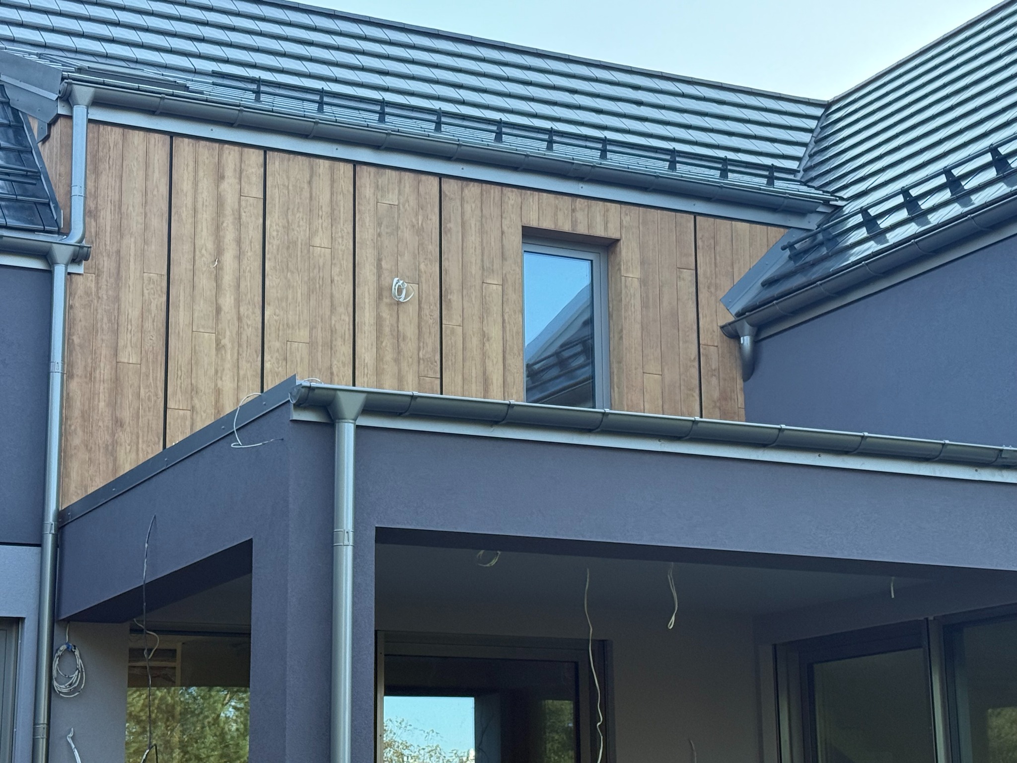 Nowoczesny dom z drewnianą elewacją i szarymi elementami. Widoczne okno, dach z dachówki i rynny. Detal wykończenia zewnętrznego domu.