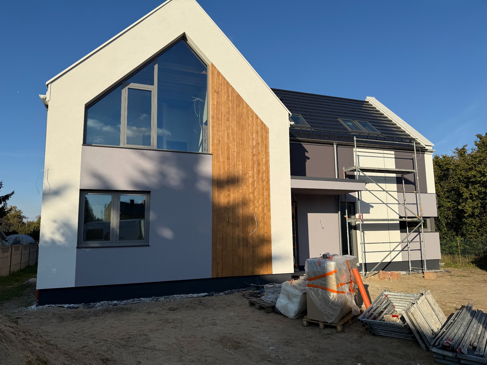 Nowoczesny dom jednorodzinny z elewacją w odcieniach szarości i beżu, z elementami drewnianymi i rusztowaniem. Widoczne materiały budowlane.