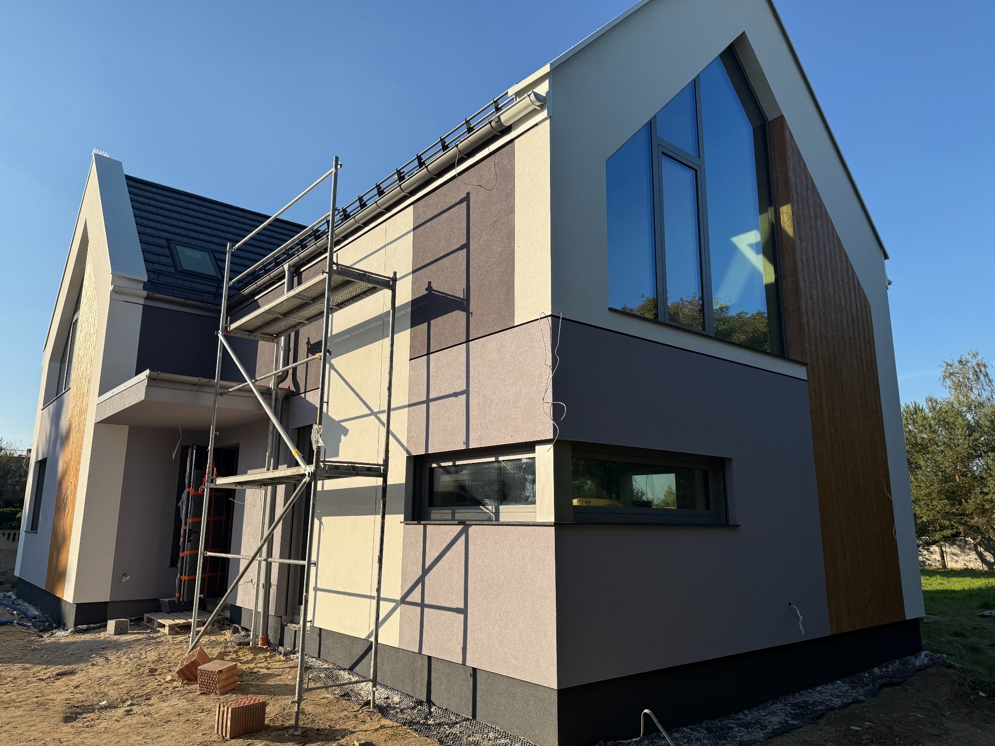 Elewacja domu w trakcie wykańczania, widoczne rusztowania. Nowoczesne detale architektoniczne, pionowe pasy w odcieniach beżu i brązu. Częściowo otynkowana.