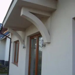 Elewacja domu z oknami i ozdobnym zadaszeniem nad wejściem, oświetlona lampami zewnętrznymi. Widoczna faktura tynku i elementy architektoniczne.