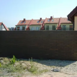 Nowoczesny mur z brązowej cegły klinkierowej na tle dom&oacute;w z czerwonymi dachami. Widoczne trawa i kamienie. Słoneczny dzień.