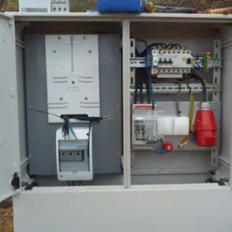 Otwarta skrzynka elektryczna z zainstalowanymi elementami: bezpieczniki, styczniki, gniazdo siłowe, przewody w różnych kolorach, zamontowane na szynach DIN.