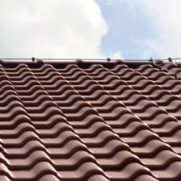 Czerwona dachówka ceramiczna z widocznym systemem odgromowym na tle błękitnego nieba z chmurami.