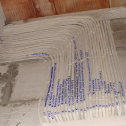 Wiązka przewodów elektrycznych ułożonych w rurkach ochronnych, przymocowanych do surowej ściany z bloczków, z odręcznymi notatkami identyfikującymi obwody.