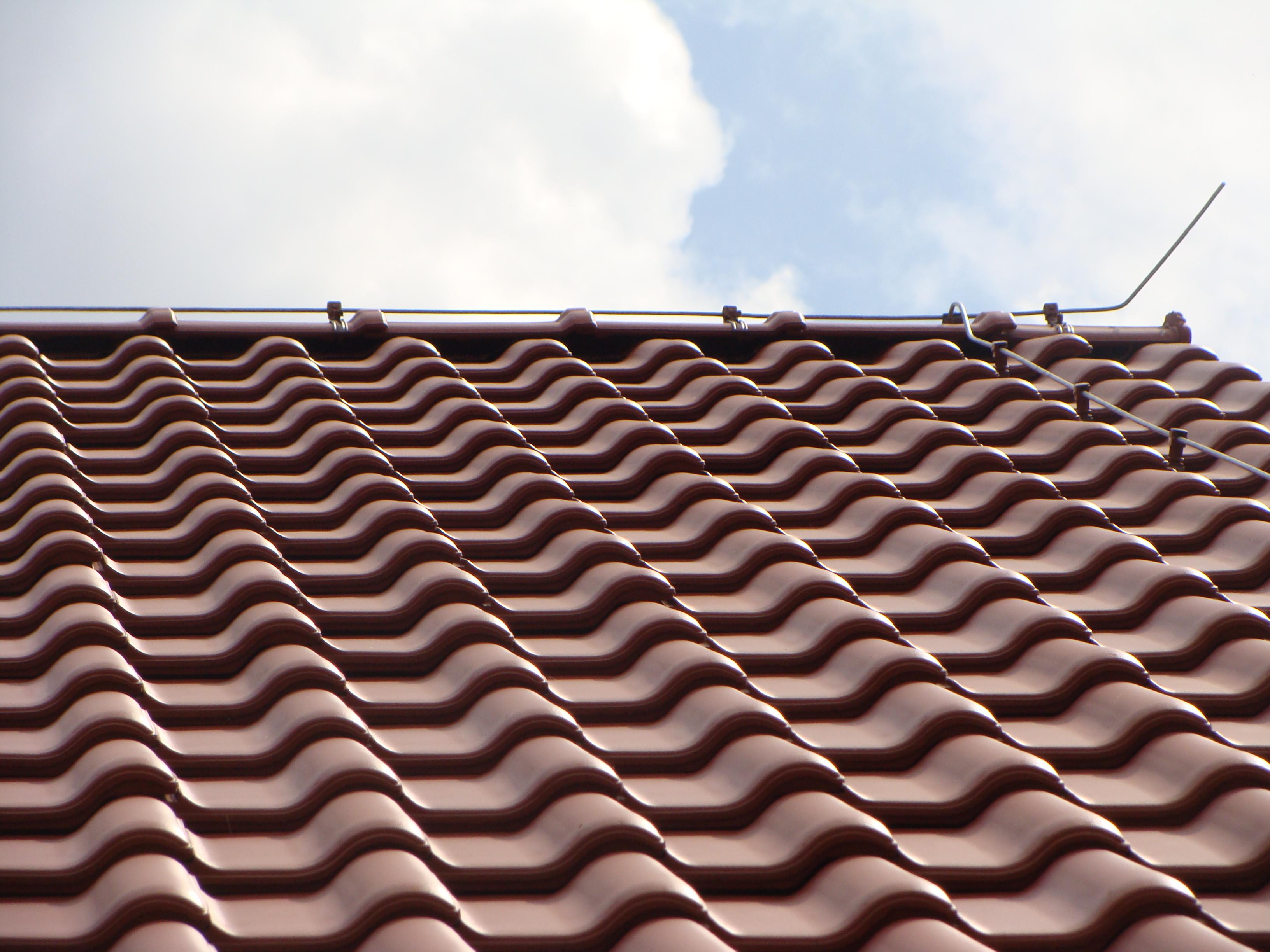 Czerwona dachówka ceramiczna z widocznym systemem odgromowym na tle błękitnego nieba z chmurami.