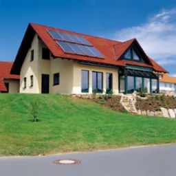 Dom jednorodzinny z czerwonym dachem, na którym zamontowane są panele słoneczne, z przeszkloną werandą i zielonym trawnikiem.