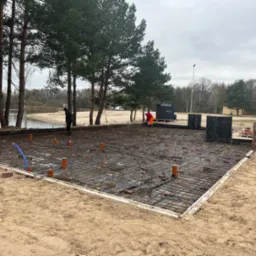 Przygotowanie zbrojenia pod fundament płyty w Krzyżanowicach, widoczne rury kanalizacyjne i pracownicy na tle jeziora.