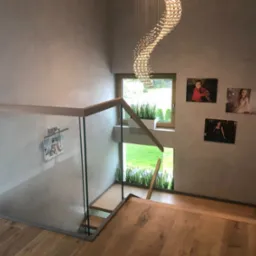 Nowoczesne schody drewniane z balustradą szklaną, oświetlone dekoracyjnym żyrandolem, z widokiem na zieleń przez okno i portretami na ścianie.