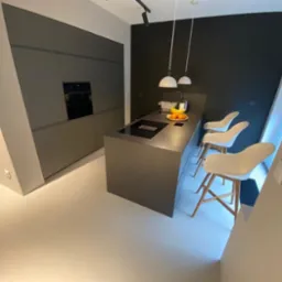 Nowoczesna kuchnia z szarymi szafkami i wyspą z szarym blatem, na której stoją owoce, oświetlona trzema wiszącymi lampami i trzema białymi krzesłami barowymi.