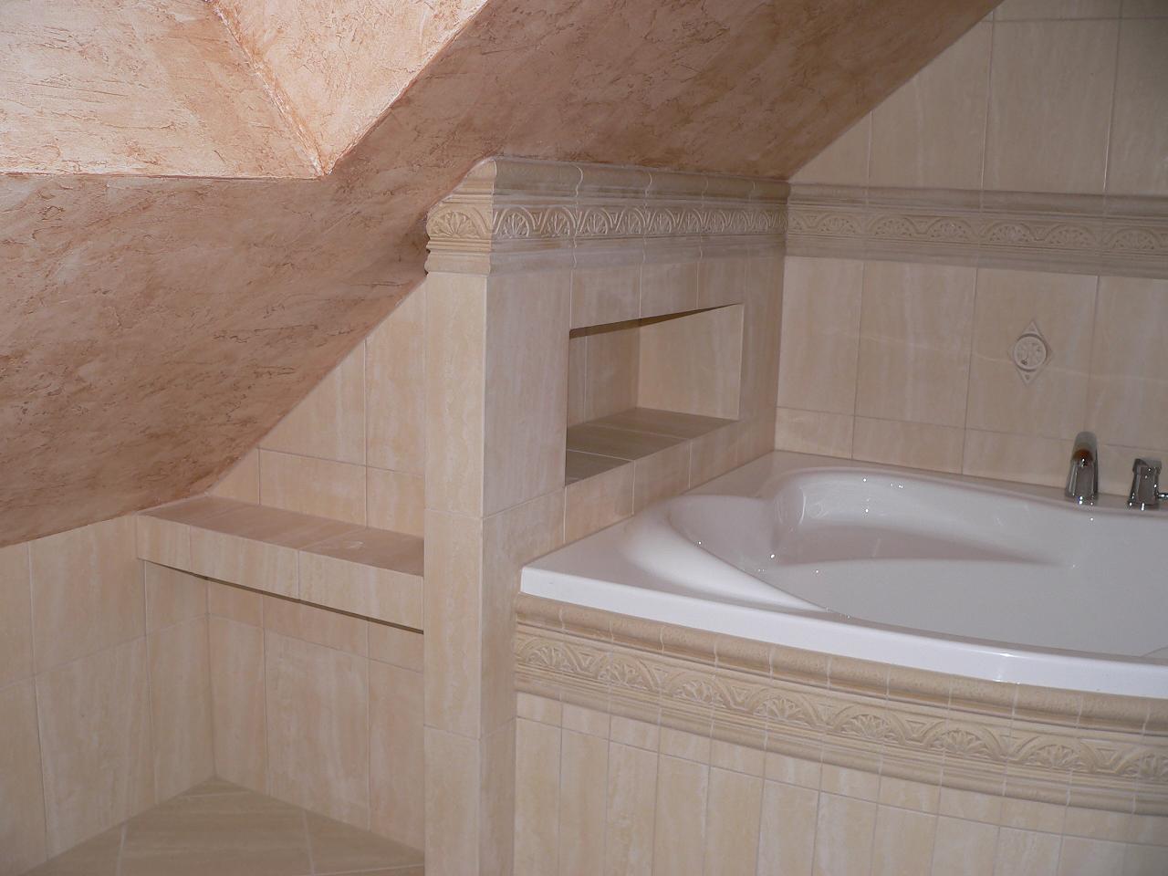 Wykończona łazienka na poddaszu z beżowymi płytkami, zabudowaną wanną, półką w ścianie i ozdobną listwą.