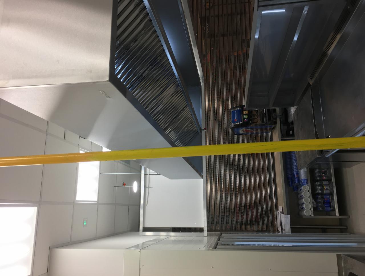 Wnętrze kuchni przemysłowej z widocznymi systemami wentylacyjnymi, instalacją elektryczną i regałem z napojami.