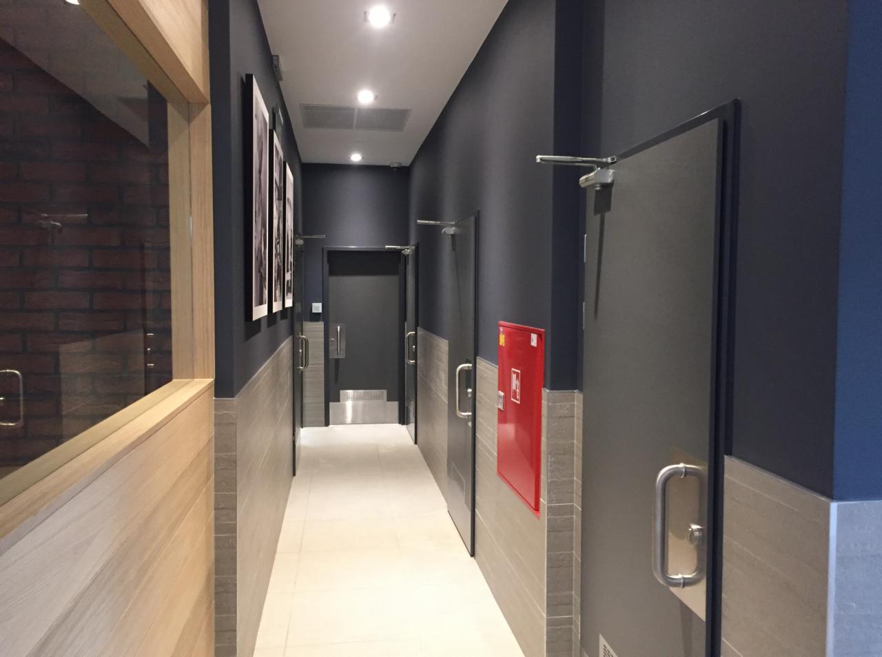 Wnętrze korytarza biurowego z szarymi drzwiami, czerwonym hydrantem i jasną podłogą z płytek, widok z perspektywy, z elementami drewnianej boazerii i obrazami na ścianie.