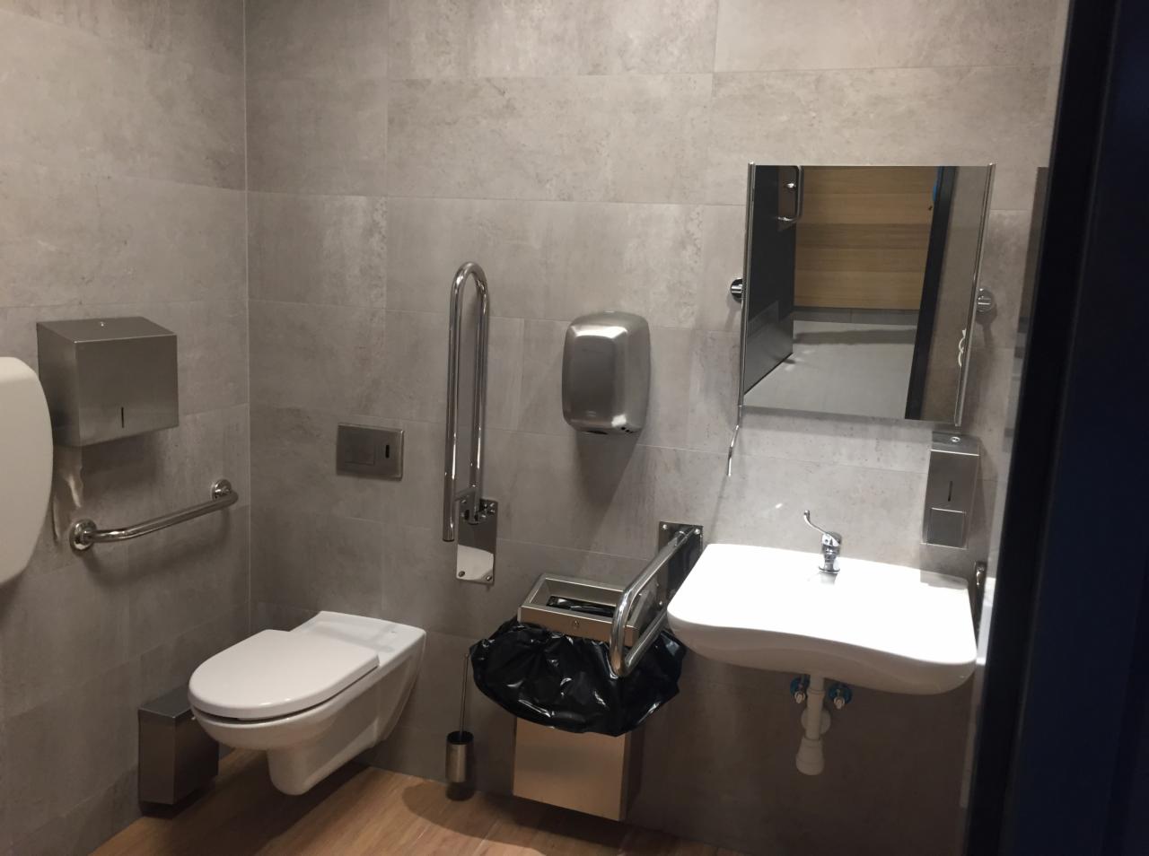 Toaleta publiczna z szarymi płytkami, podwieszaną miską WC, umywalką, lustrem i uchwytami dla niepełnosprawnych, widok z niskiego kąta.
