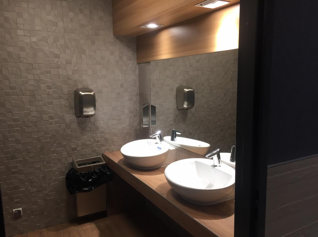 Nowoczesna łazienka publiczna z dwoma białymi umywalkami nablatowymi na drewnianym blacie, szara mozaika na ścianach, automatyczne suszarki do rąk oraz kosz na śmieci z czarnym workiem.