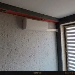 Biały klimatyzator marki Gree zamontowany na ścianie z dekoracyjnej, jasnej cegły w pomieszczeniu z oknem przesłoniętym roletami.