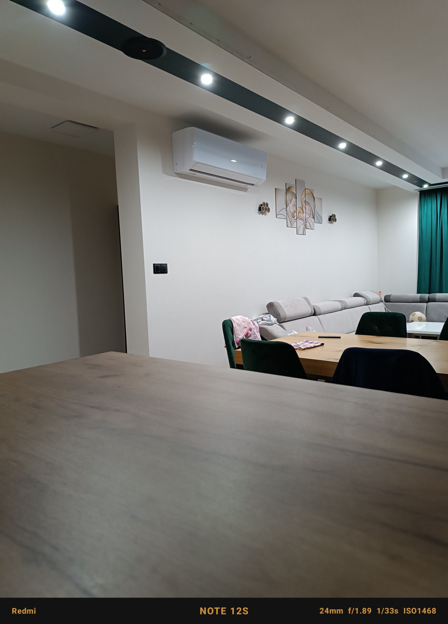 Biały klimatyzator ścienny zamontowany w nowoczesnym salonie z szarą sofą, ciemnozielonymi krzesłami i dekoracyjnymi obrazami na ścianie.