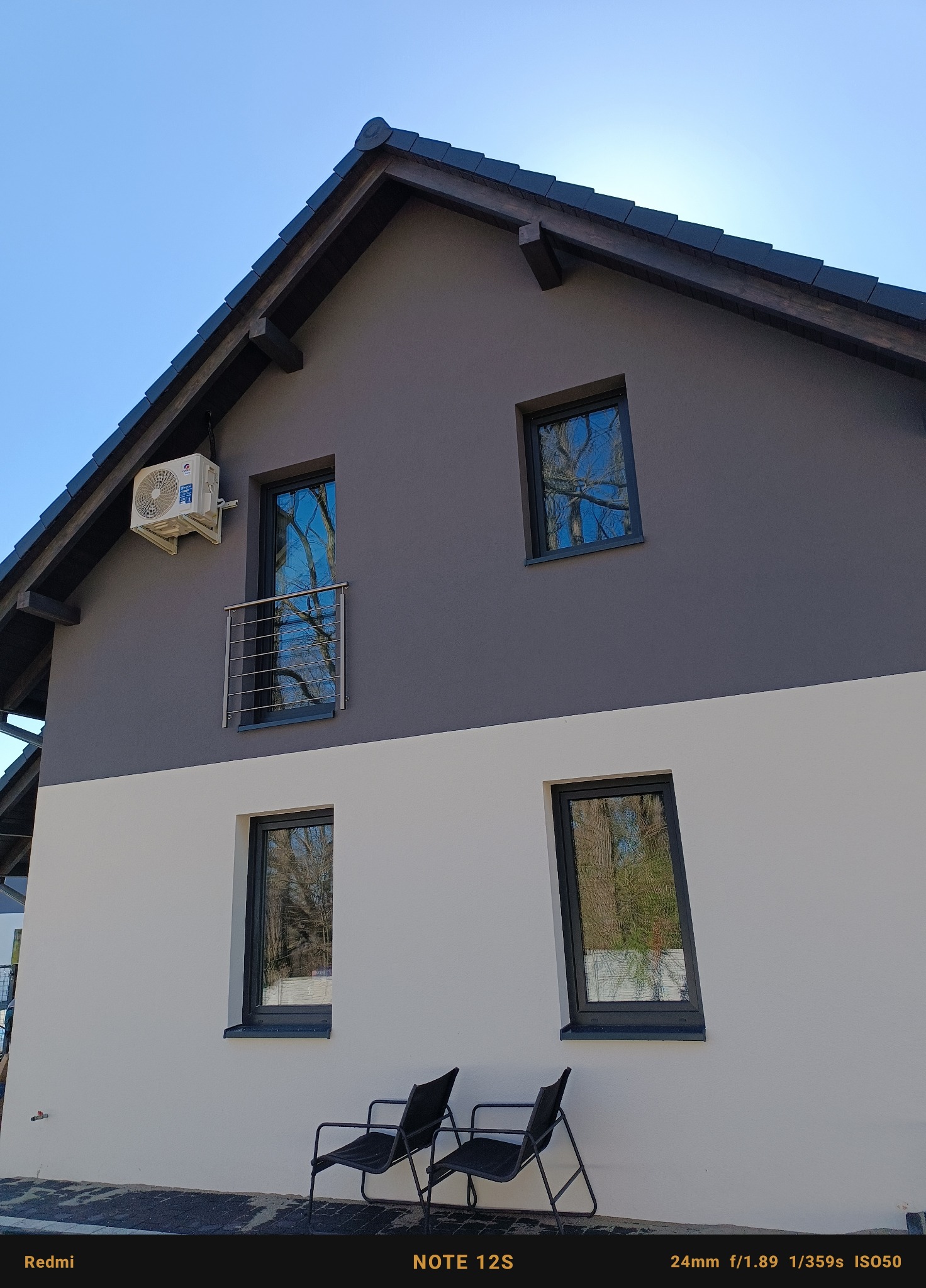 Dwukondygnacyjny dom z zainstalowaną jednostką zewnętrzną klimatyzacji na szarej ścianie, widok z dołu na błękitne niebo, dwa krzesła ogrodowe przed białą ścianą.