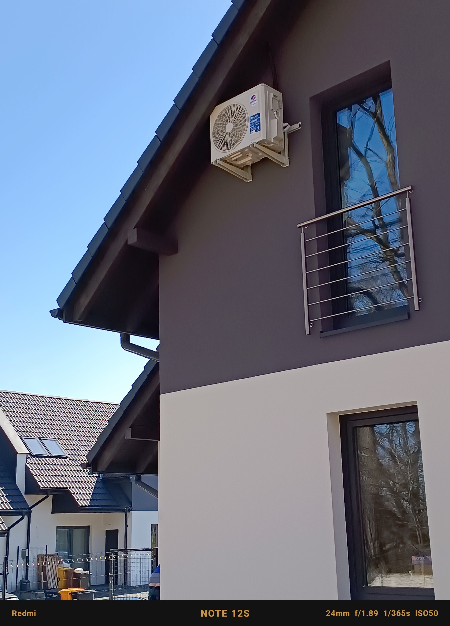 Biała jednostka klimatyzacji zamontowana na szarej ścianie dwukondygnacyjnego domu z białą dolną częścią elewacji, widok z dołu na błękitne niebo.