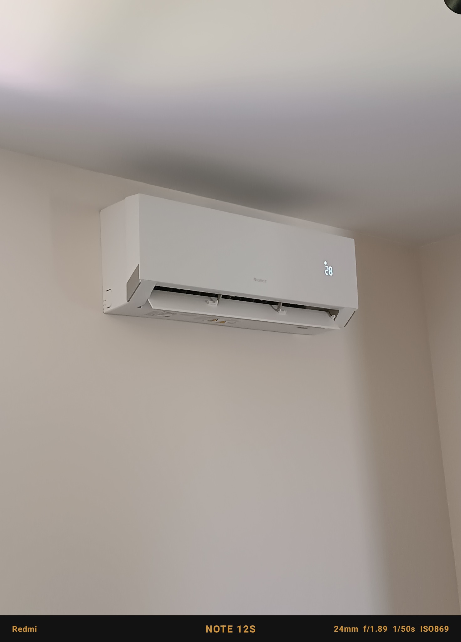 Biały klimatyzator marki Gree zamontowany na jasnej ścianie, wyświetlający temperaturę 28 stopni Celsjusza.