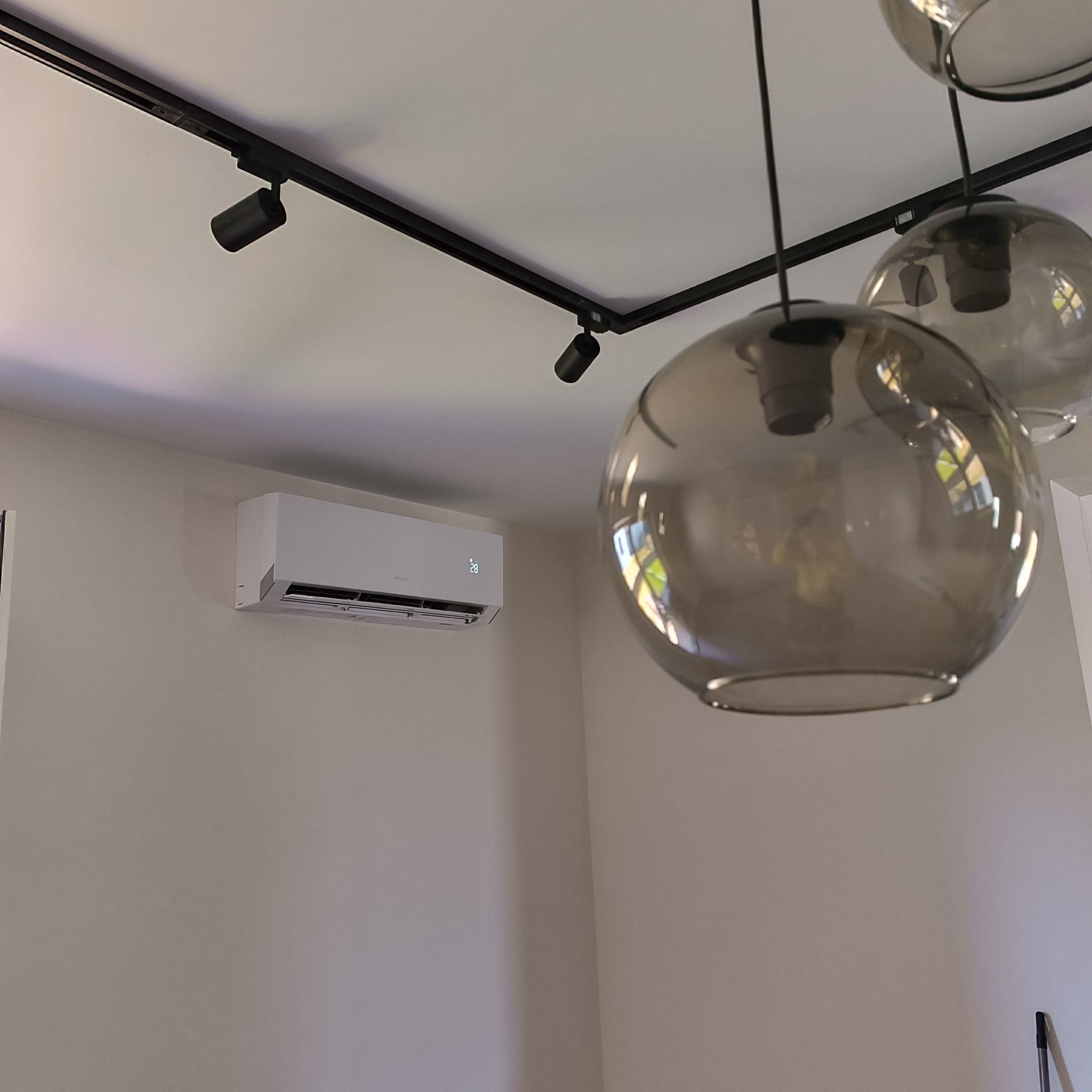 Biały klimatyzator ścienny w minimalistycznym wnętrzu z szynoprzewodem oświetleniowym i szklanymi lampami wiszącymi.