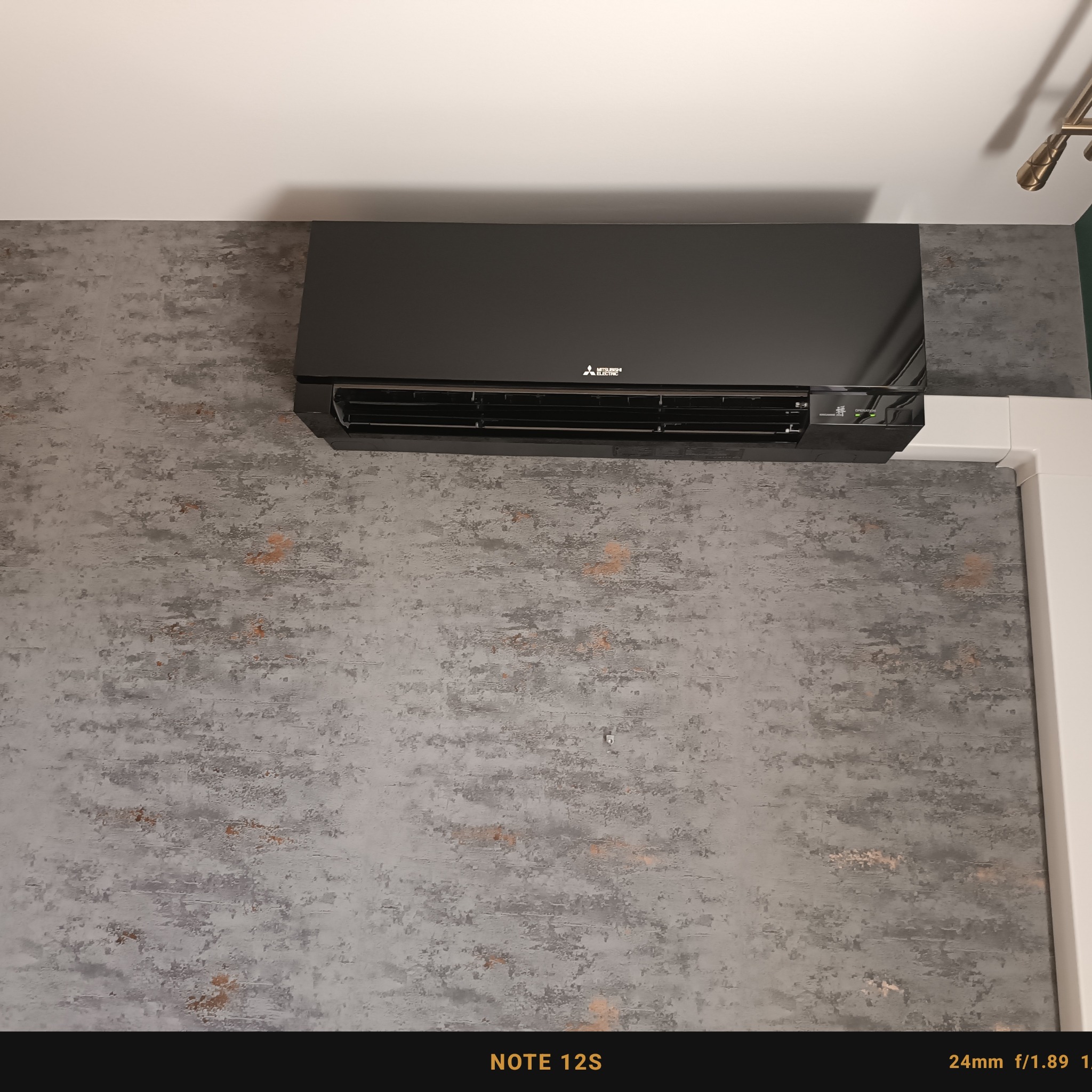 Czarna jednostka klimatyzacji Mitsubishi Electric, model NOTE 12S, zamontowana na szarej ścianie o teksturze betonu, z widocznymi białymi listwami przypodłogowymi.
