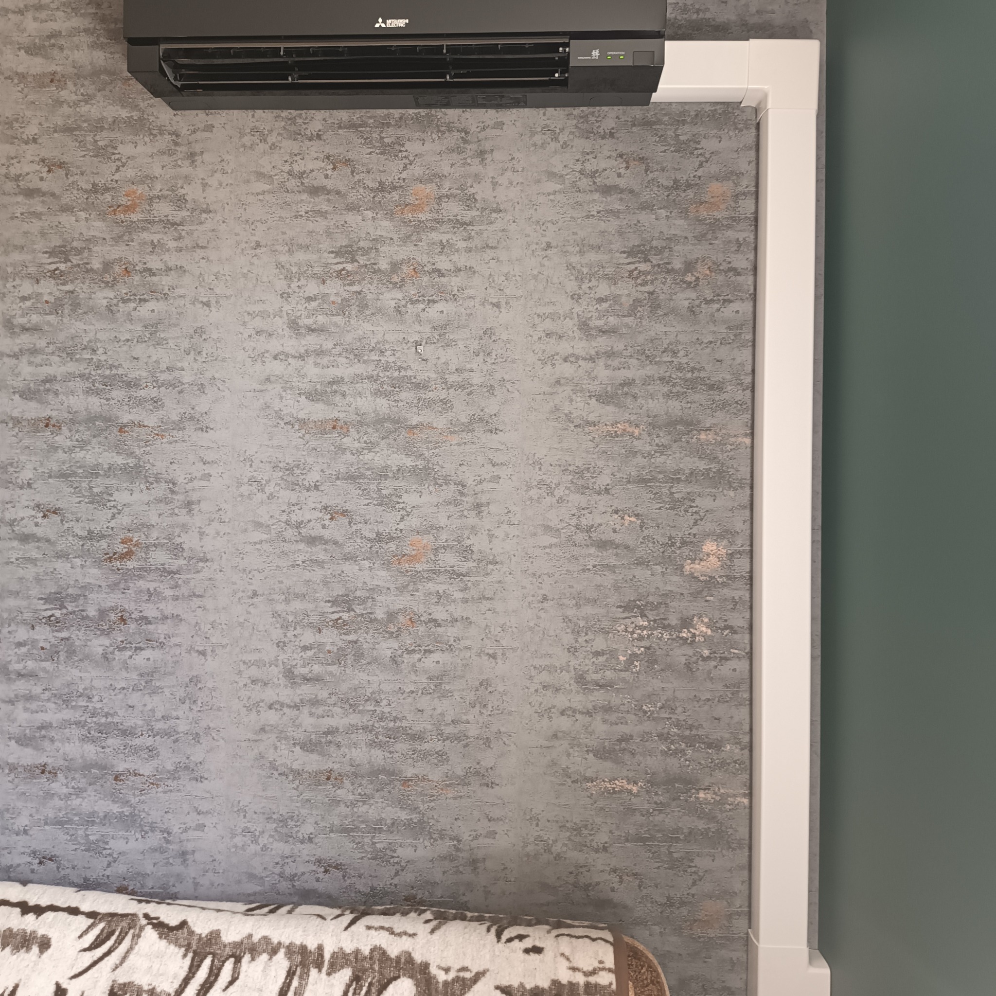 Czarny klimatyzator Mitsubishi Electric zamontowany na szarej ścianie o teksturze betonu, z widoczną białą maskownicą instalacji i fragmentem sofy w dolnej części kadru.