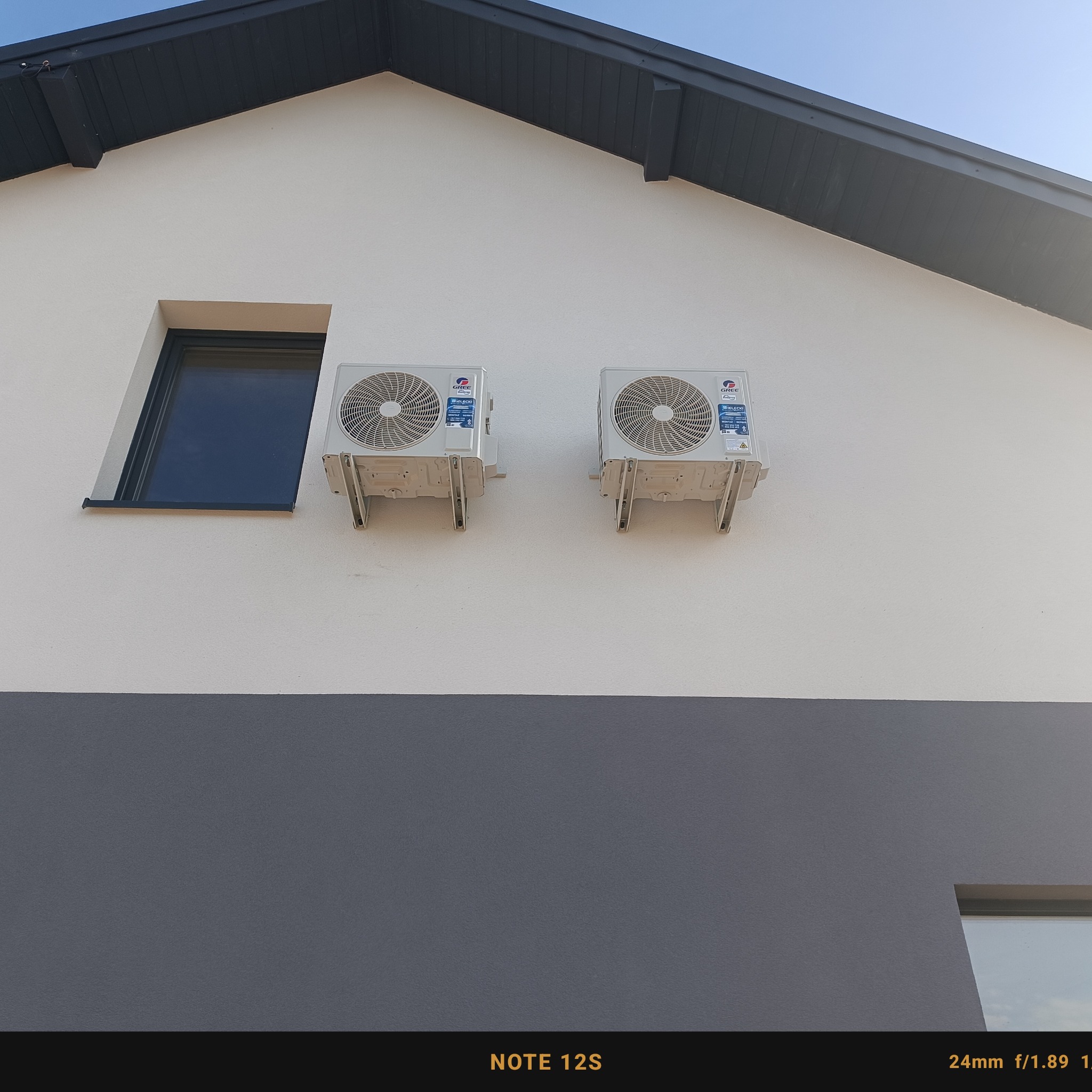 Dwa białe klimatyzatory marki Gree zamontowane na elewacji dwukondygnacyjnego domu z szarą dolną częścią i czarnym dachem, widoczne okno.