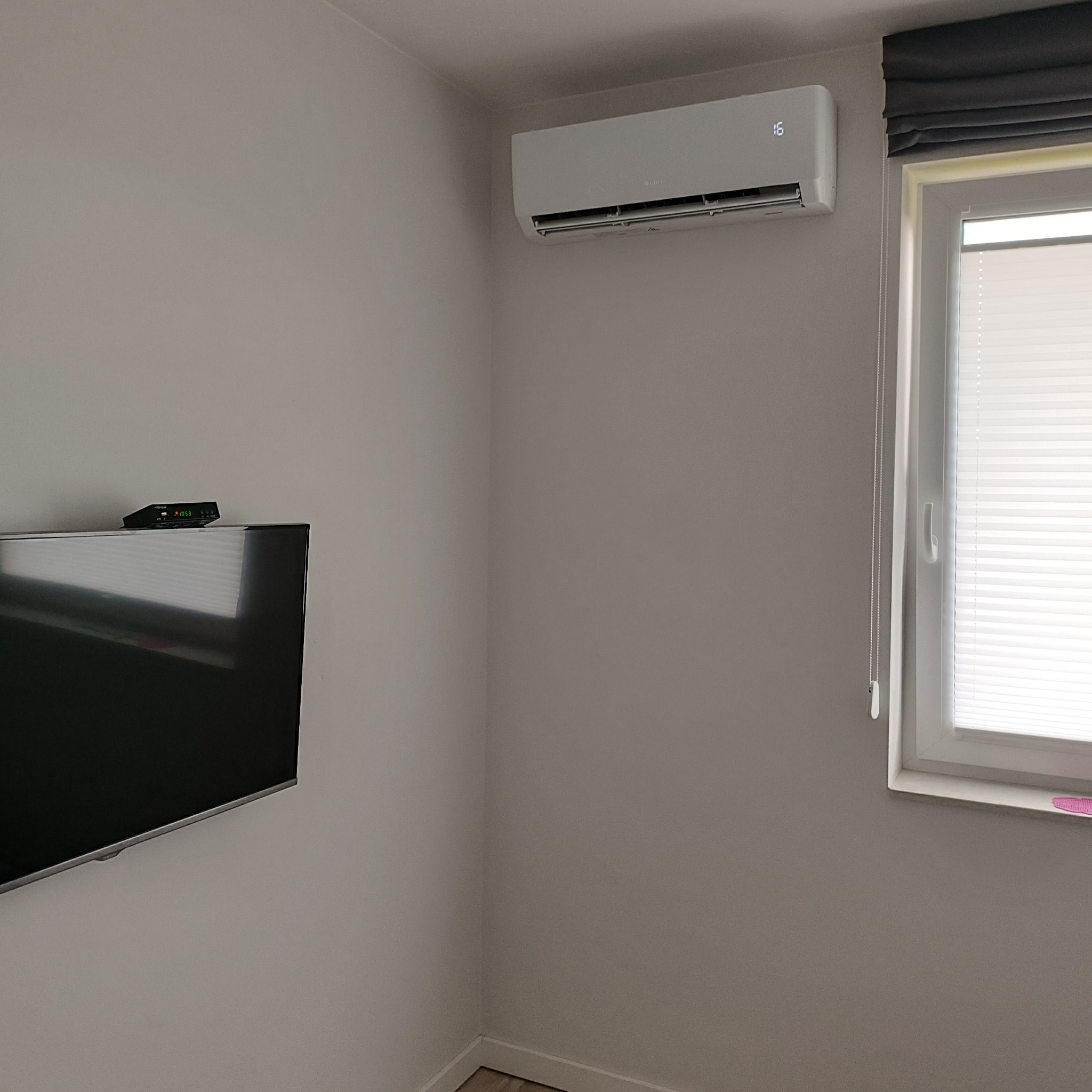 Biały klimatyzator ścienny zamontowany w rogu pokoju, widoczny wyświetlacz temperatury '16', obok okno z roletami plisowanymi i zasłoną rzymską, na ścianie telewizor.